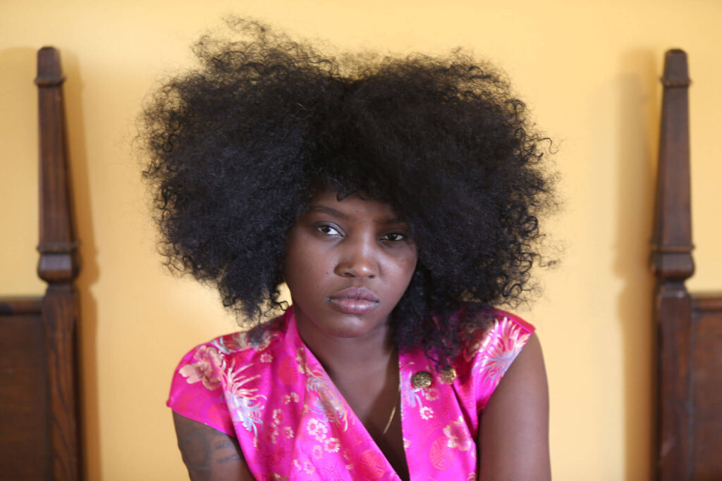 Portrait of a woman with natural curly hair, wearing a vibrant pink floral top against a yellow background.