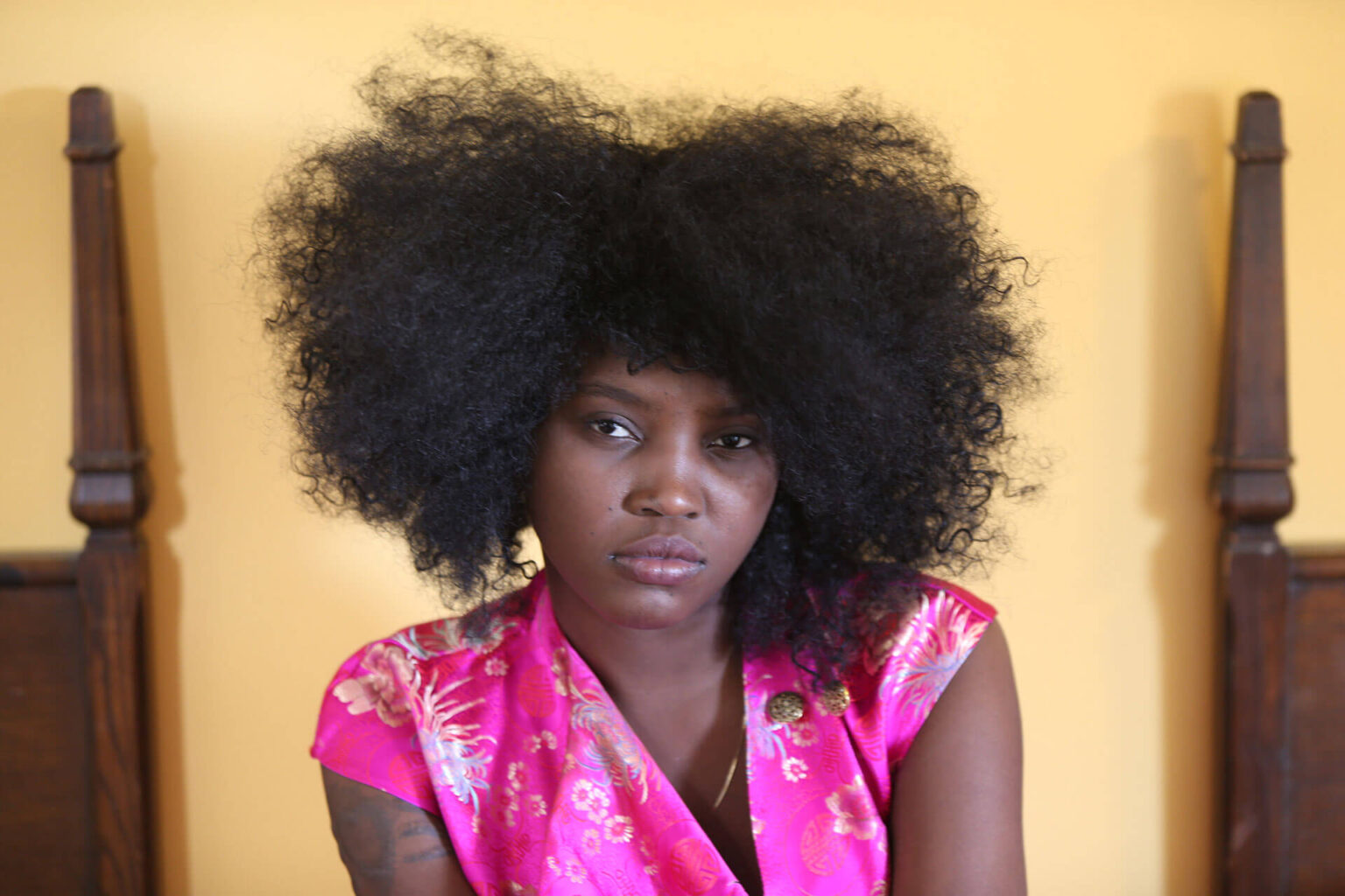 Portrait of a woman with natural curly hair, wearing a vibrant pink floral top against a yellow background.